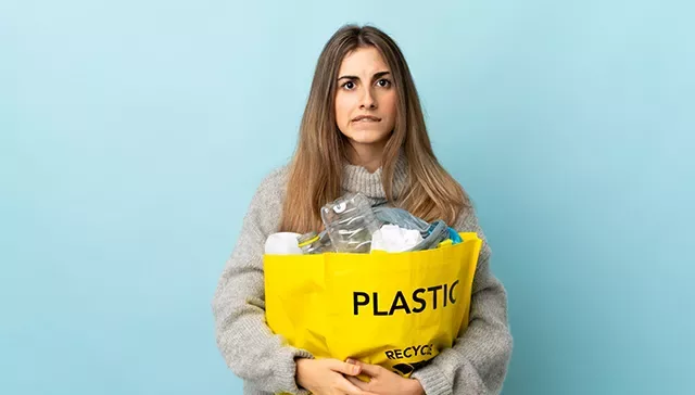 A confused looking woman holding a bag full of different types of plastic.